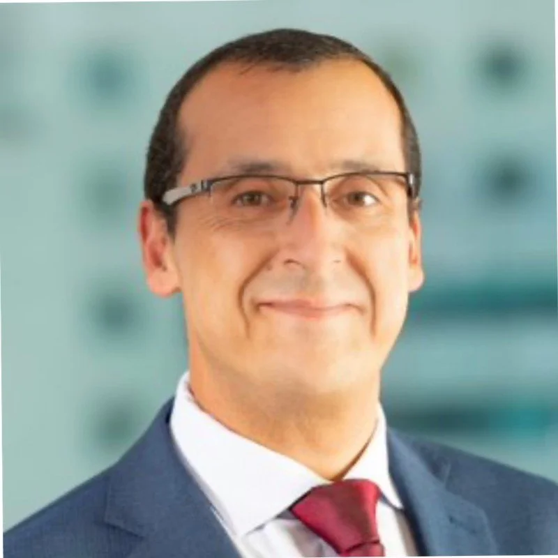 A professional man wearing glasses, a suit, and a red tie, smiling with a blurry background.