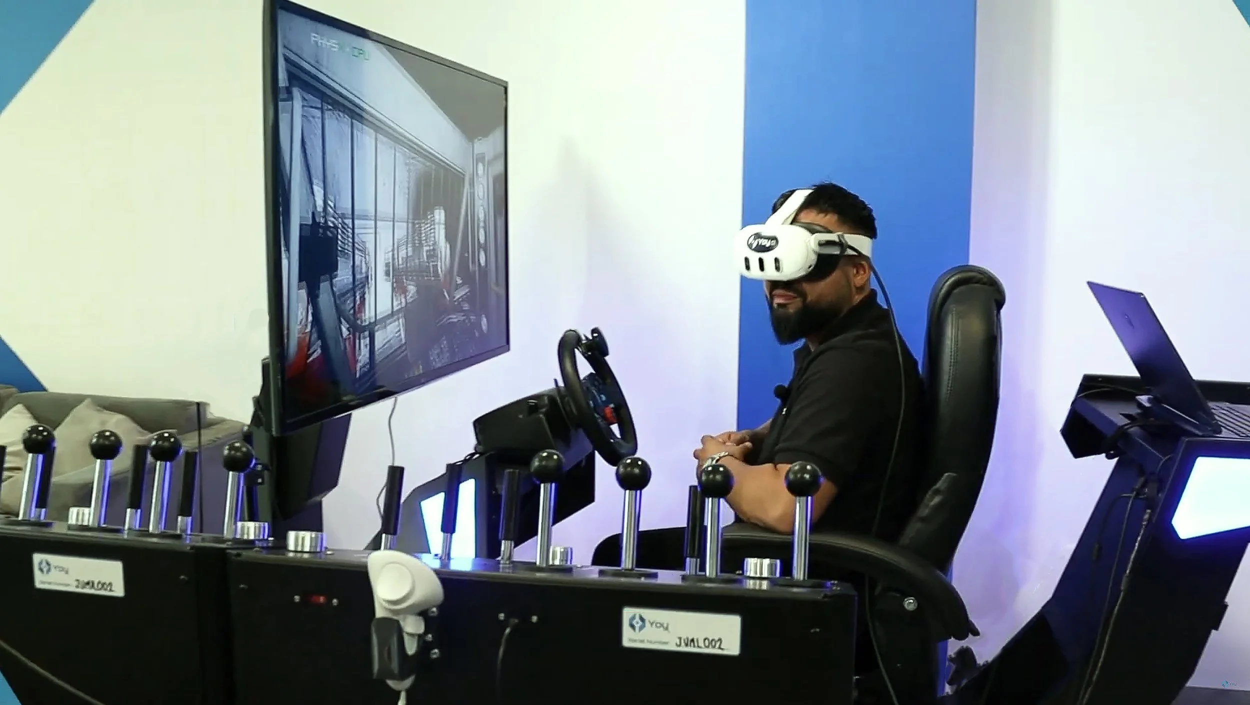 Man wearing a virtual reality headset sitting at a gaming simulator with a large screen in front of him and a racing wheel and pedals setup around him.