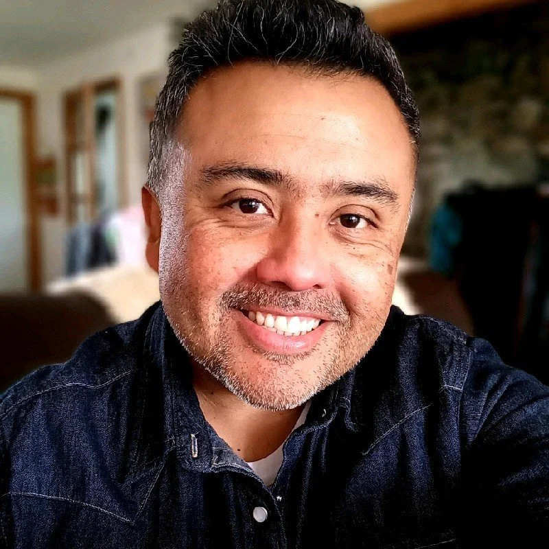 Smiling man in denim shirt indoors with blurred background.