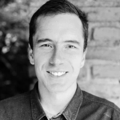 Black and white portrait of a smiling man in a collared shirt, with a brick wall background.