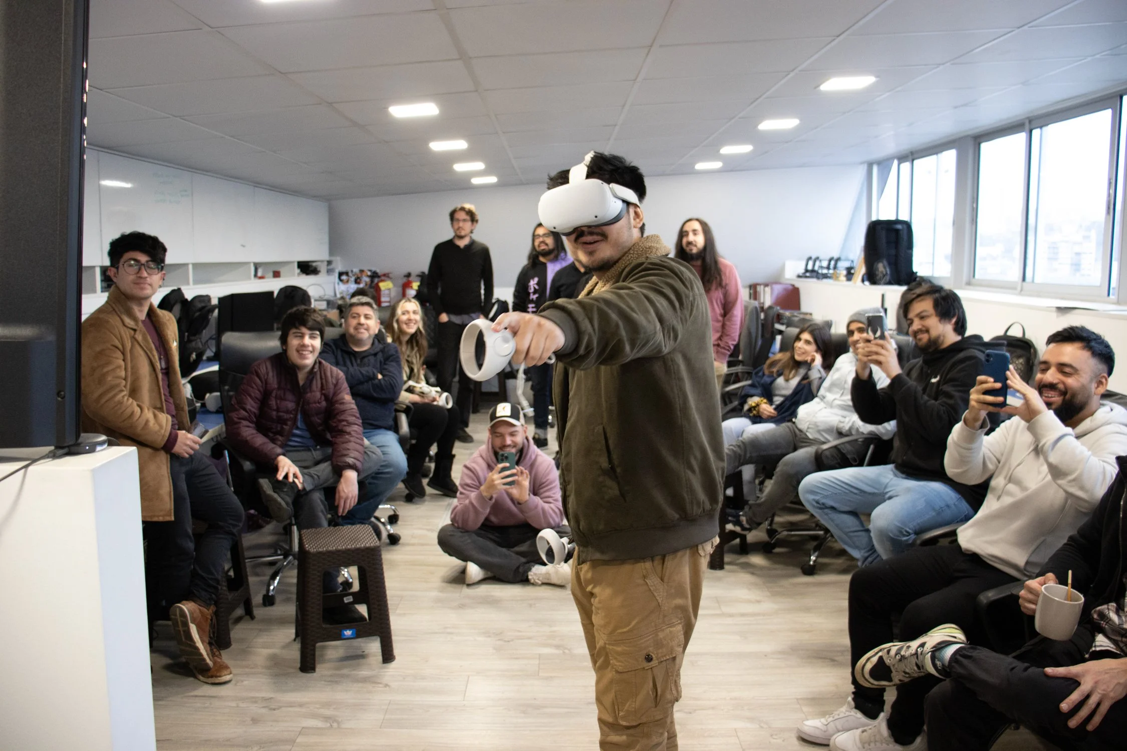 A group of people in an office setting, with one person using a virtual reality headset and controllers. Others are seated around, some taking photos with their phones, smiling and watching the VR demonstration.