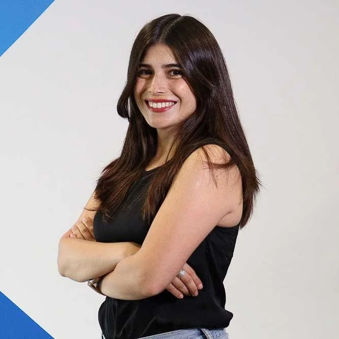 Romina Bobadilla, with long hair wearing a black sleeveless top, arms crossed, standing against a light background with blue corner accents.