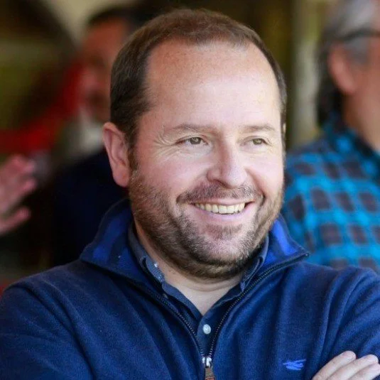 Smiling man wearing a blue jacket with plaid shirt, standing indoors with another person blurred in the background.