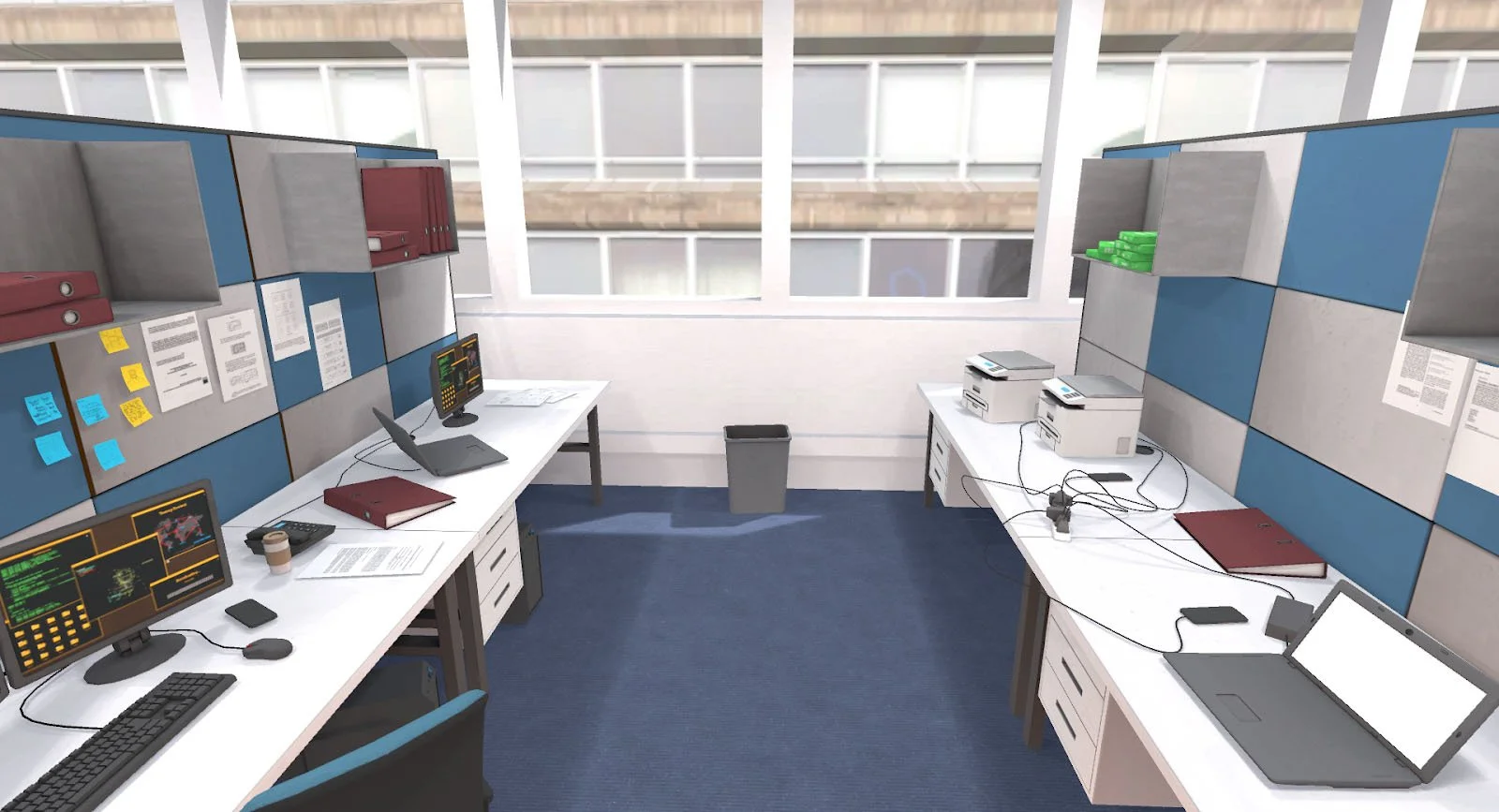 Interior of an office space with two desks, each equipped with laptops and monitors. One desk has a folder, coffee cup, and documents, while the other has cables, printers, and more folders. The workspace includes blue and gray partition walls with shelves and windows in the background.