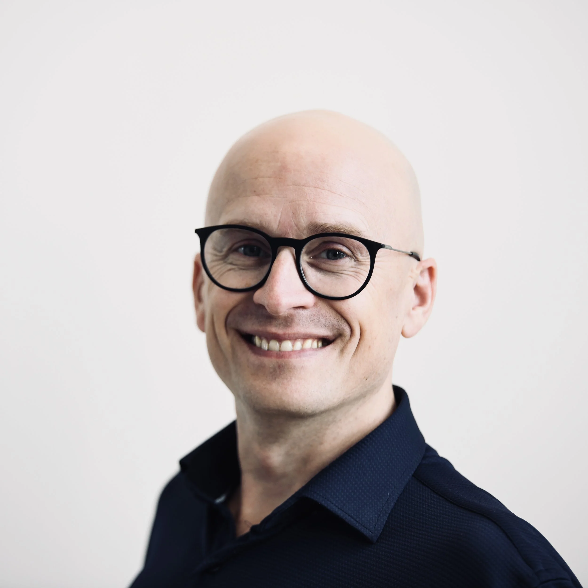 Alistair Schneider wearing black glasses and a black shirt smiling in front of a white background.