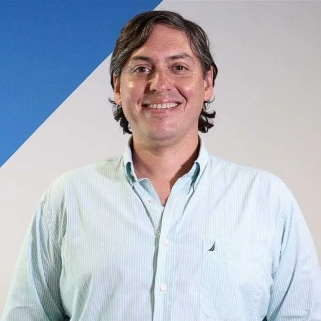 Tomás Chales in a light blue button-up shirt standing against a white and blue geometric background, smiling at the camera.