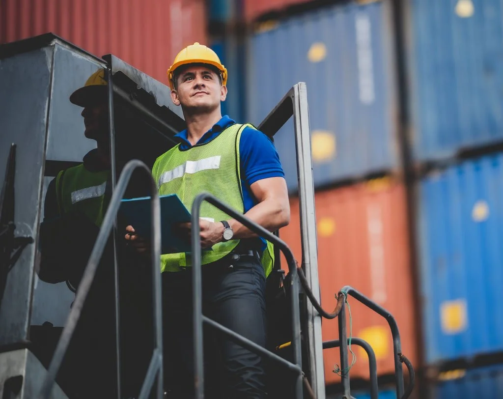 Construction worker in a hard hat and safety vest near shipping containers, holding a clipboard.