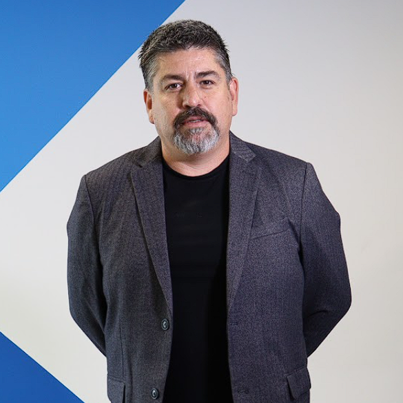 Emerson Puelles with a gray beard in a suit standing against a geometric blue and white background.