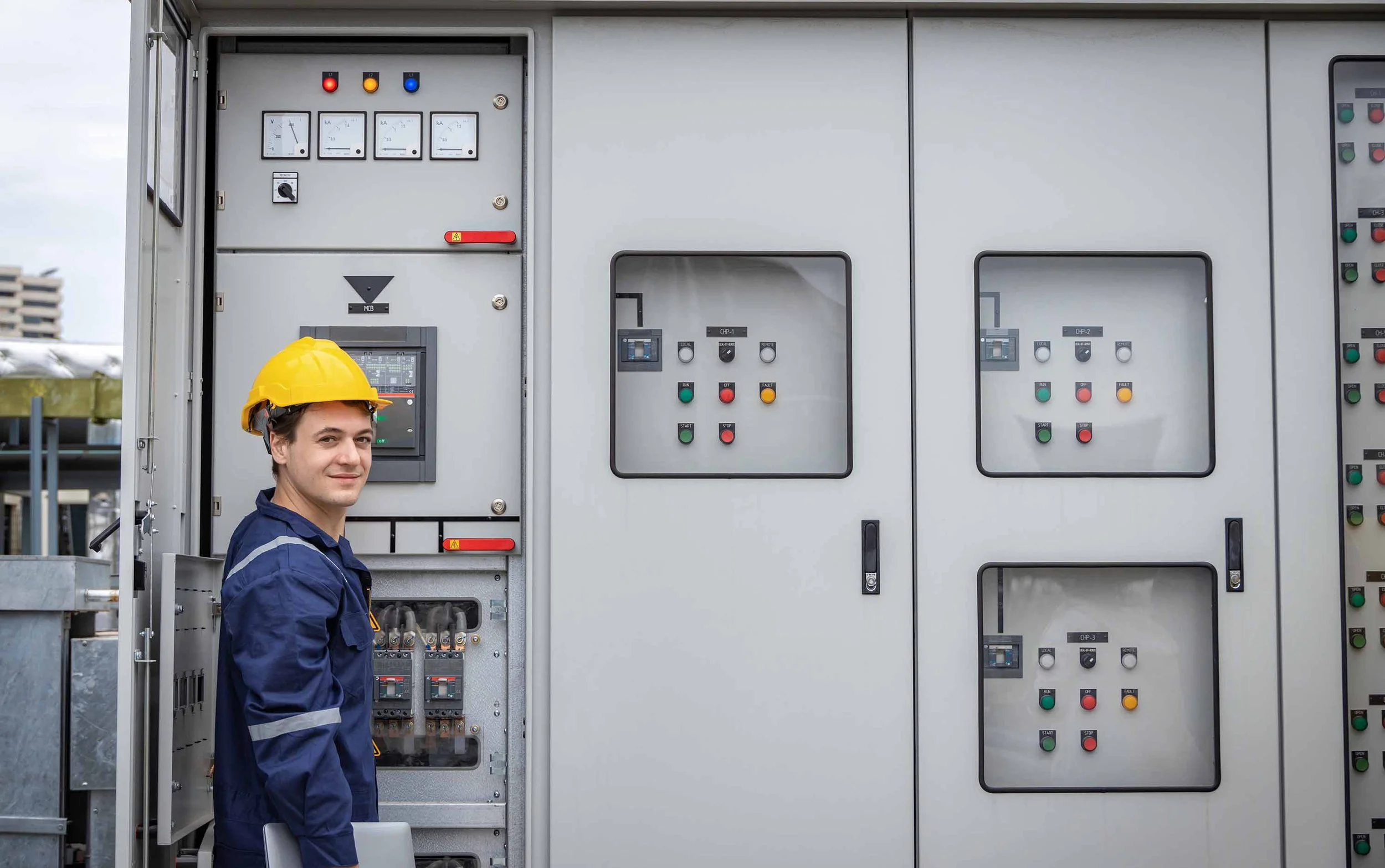 A worker in safety gear standing next to an open electrical control panel outdoors, wearing a yellow hard hat and protective clothing.